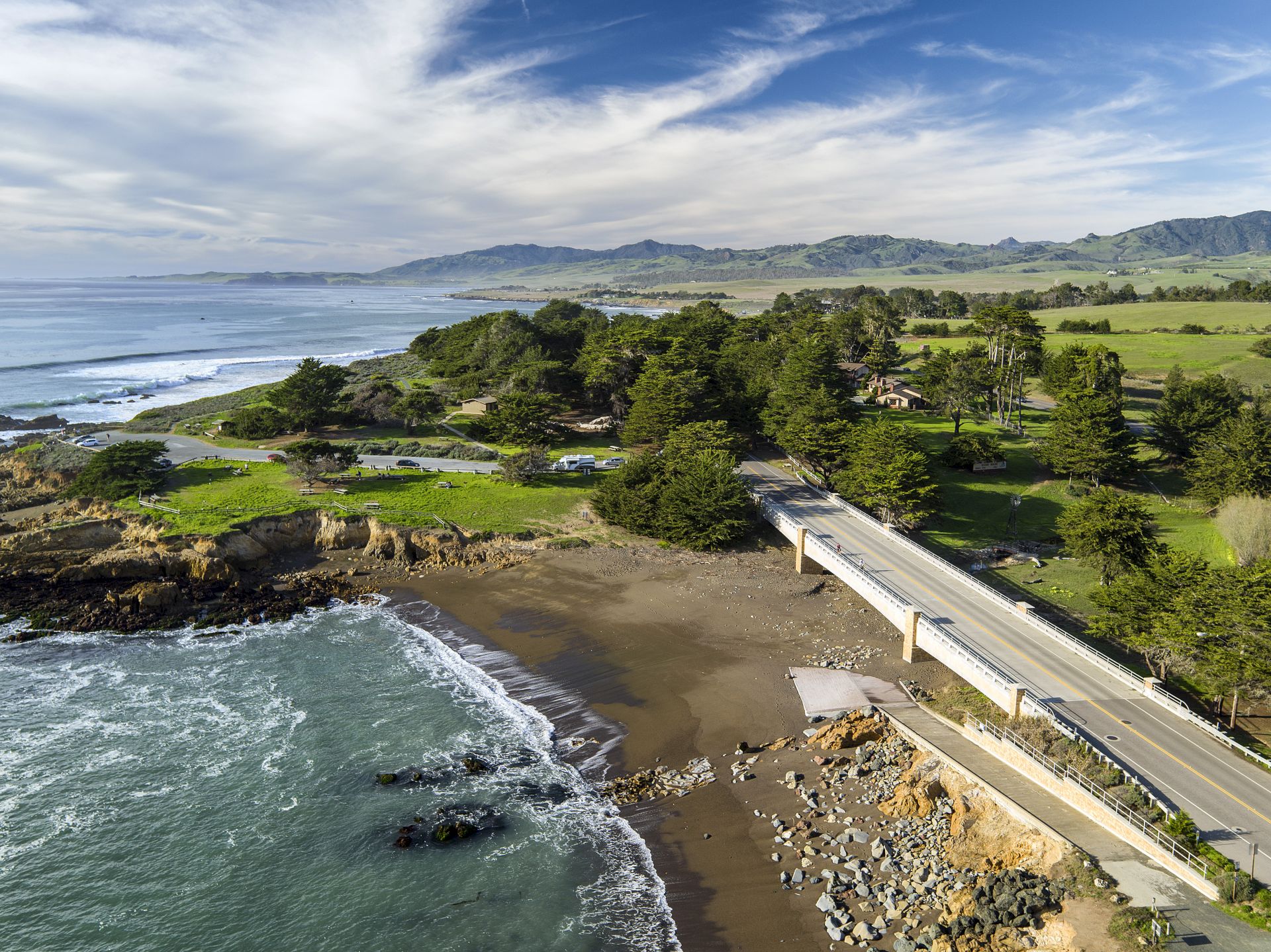 A coastal landscape with waves, sandy beach, green hills, and a bridge-like road winding through trees by the sea.
