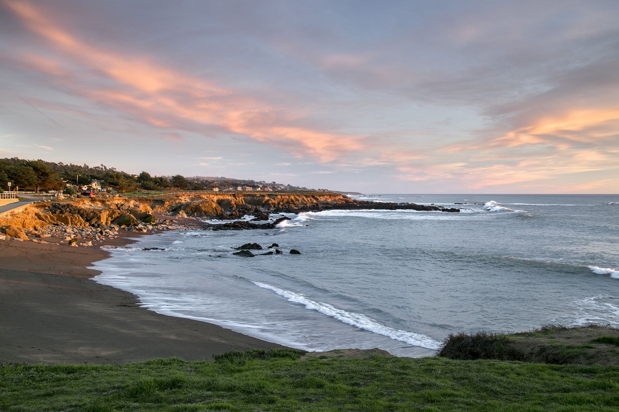 A tranquil rocky coastline at sunset, with waves rolling onto a sandy beach and warm pink clouds in the sky.