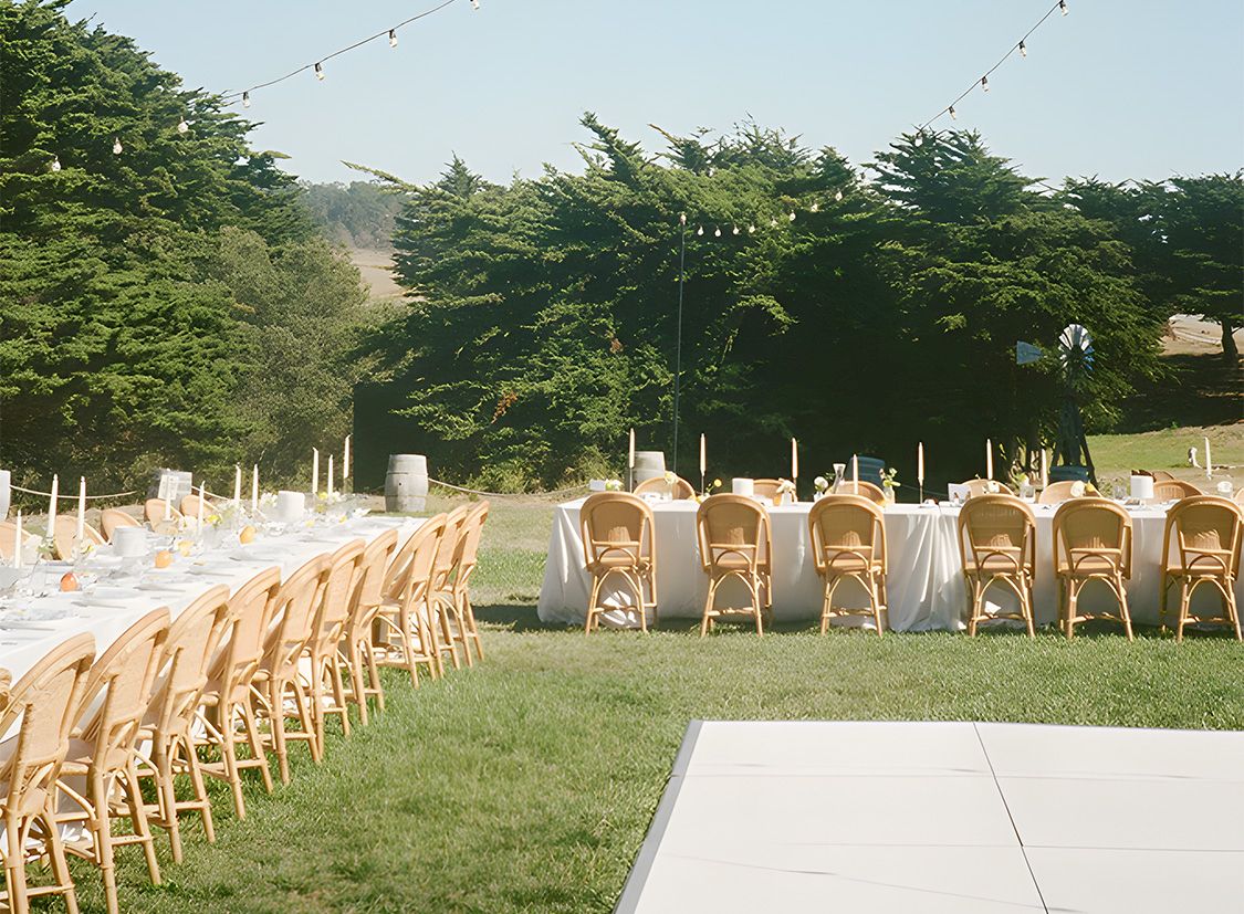 Outdoor wedding reception setup with long tables, white linens, and wooden chairs on a grassy lawn under string lights.