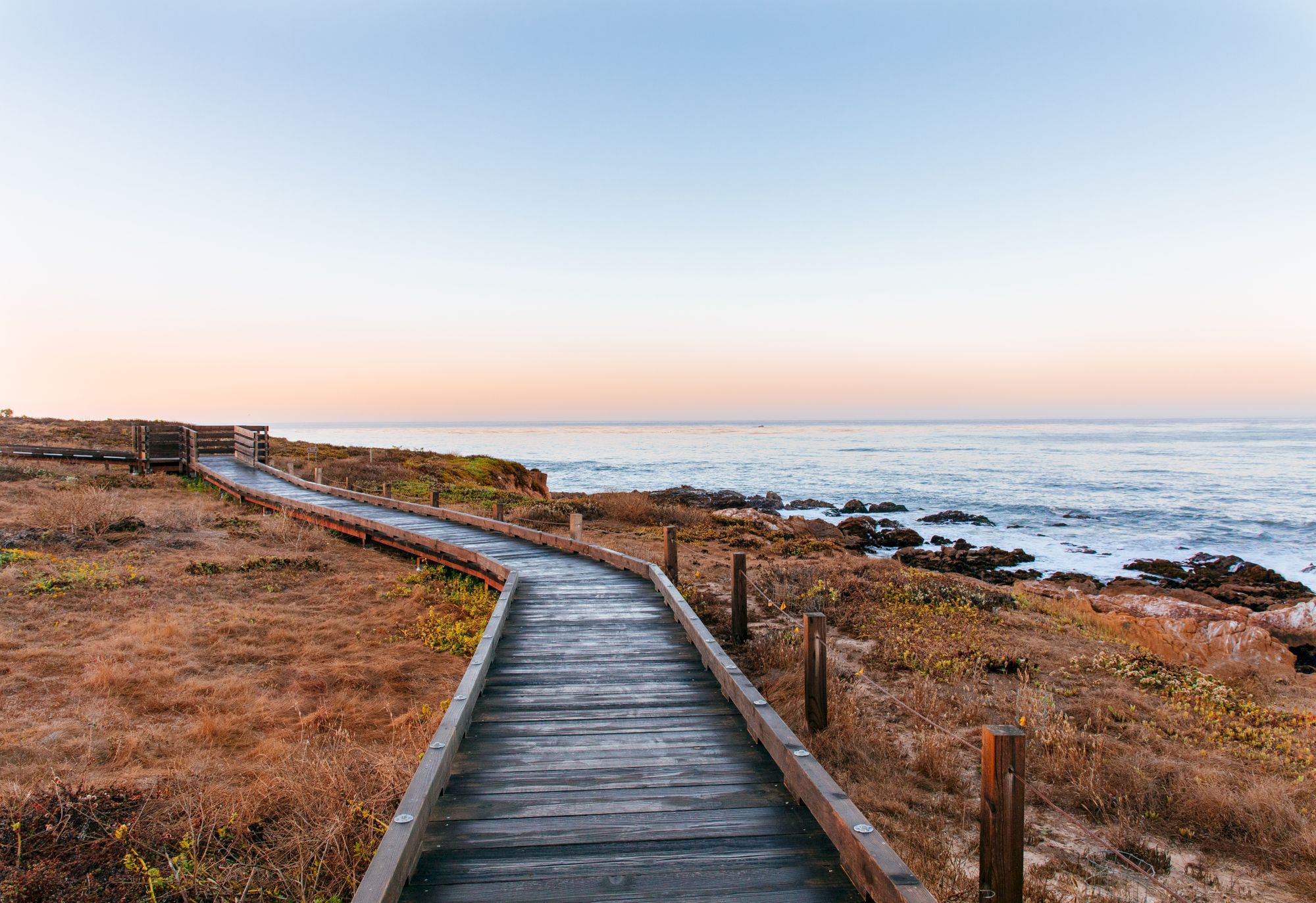 A wooden boardwalk winds along a rugged coastline at sunset, with ocean waves and rocks beside it.
