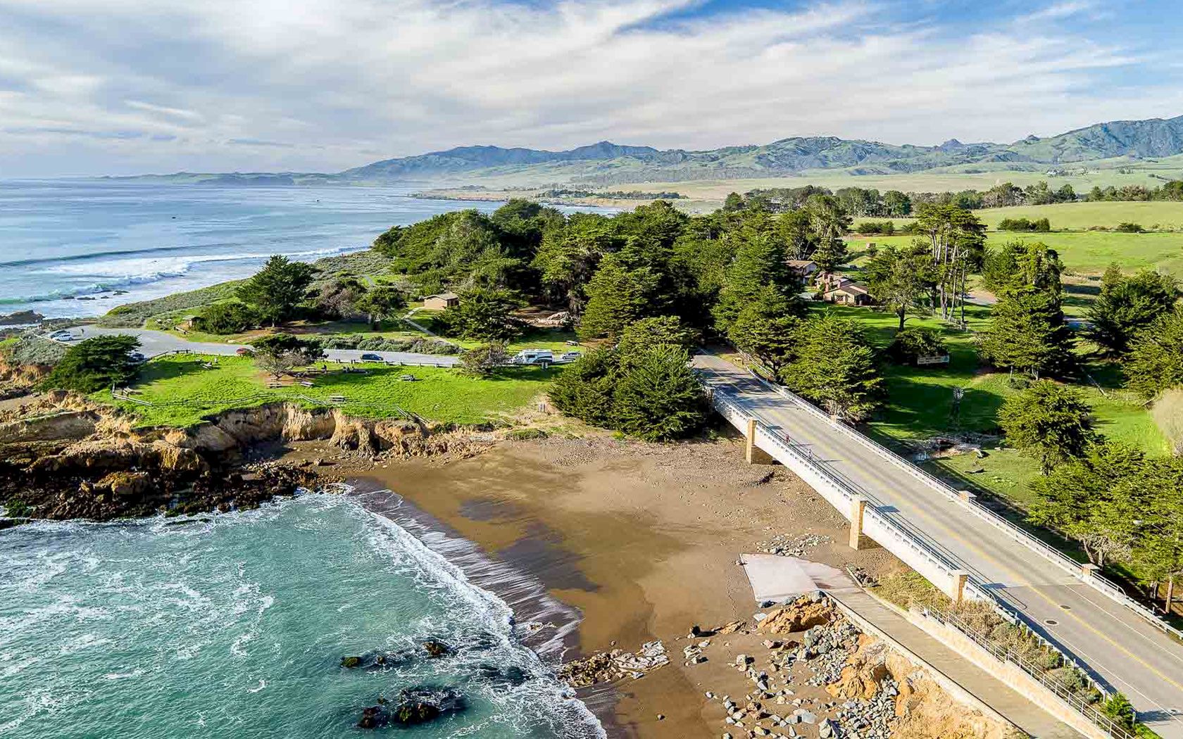A coastal bridge crosses a rocky shoreline to a sandy beach, with pine trees, green fields, and distant hills along the ocean.