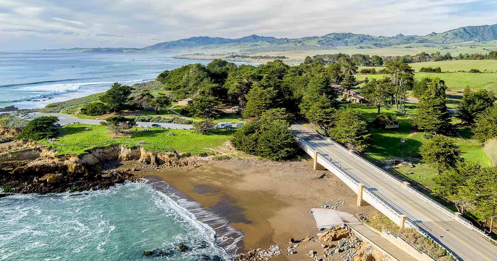 A coastal bridge crosses a rocky shoreline to a sandy beach, with pine trees, green fields, and distant hills along the ocean.