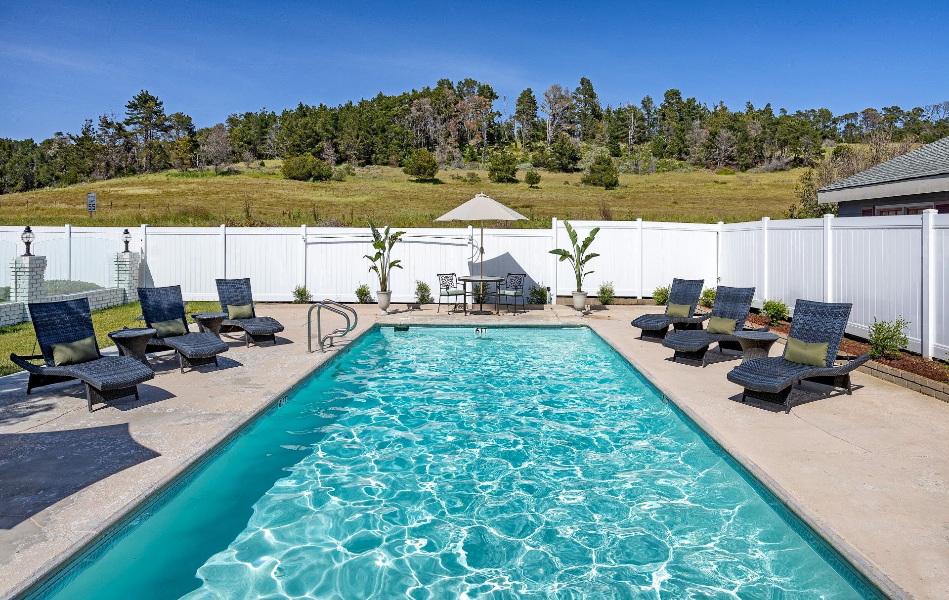 A sunny backyard pool scene with a long rectangular pool, blue water, surrounded by lounge chairs and a white fence, under a clear sky.