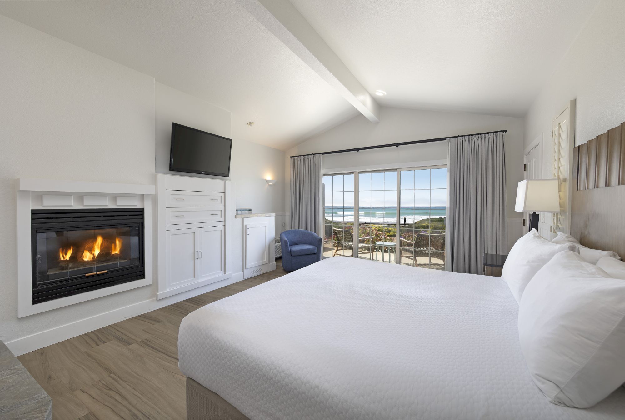 A bright beachfront bedroom with a lit fireplace, wall TV, and sliding doors showing the ocean view outside.