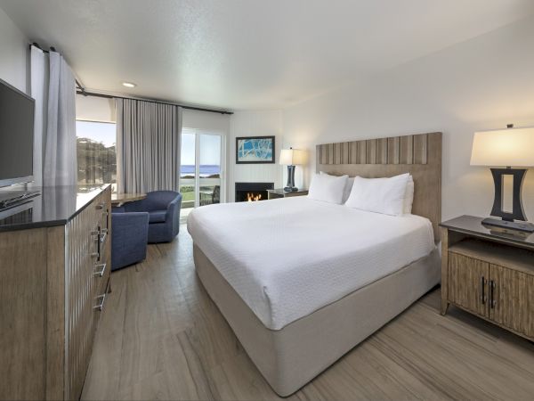 A bright hotel room with a large bed, nightstands, a flat-screen TV, a dresser, a seating area by the window, and a view toward a balcony outside.
