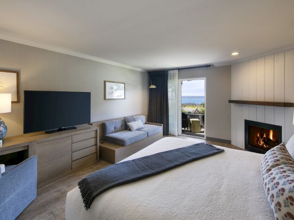 A modern hotel room with a bed, sofa, desk, TV, dresser, fireplace, and a balcony view to the outdoors.
