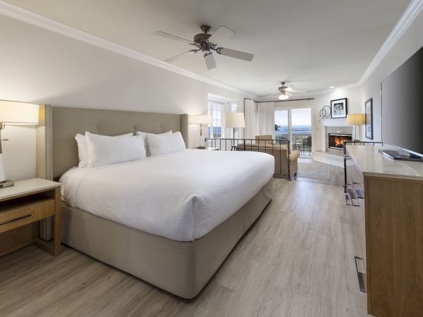A modern hotel room with a large king bed, nightstands, a desk, a flat-screen TV, light wood floors, ceiling fans, and seating by the window, tidy and bright.