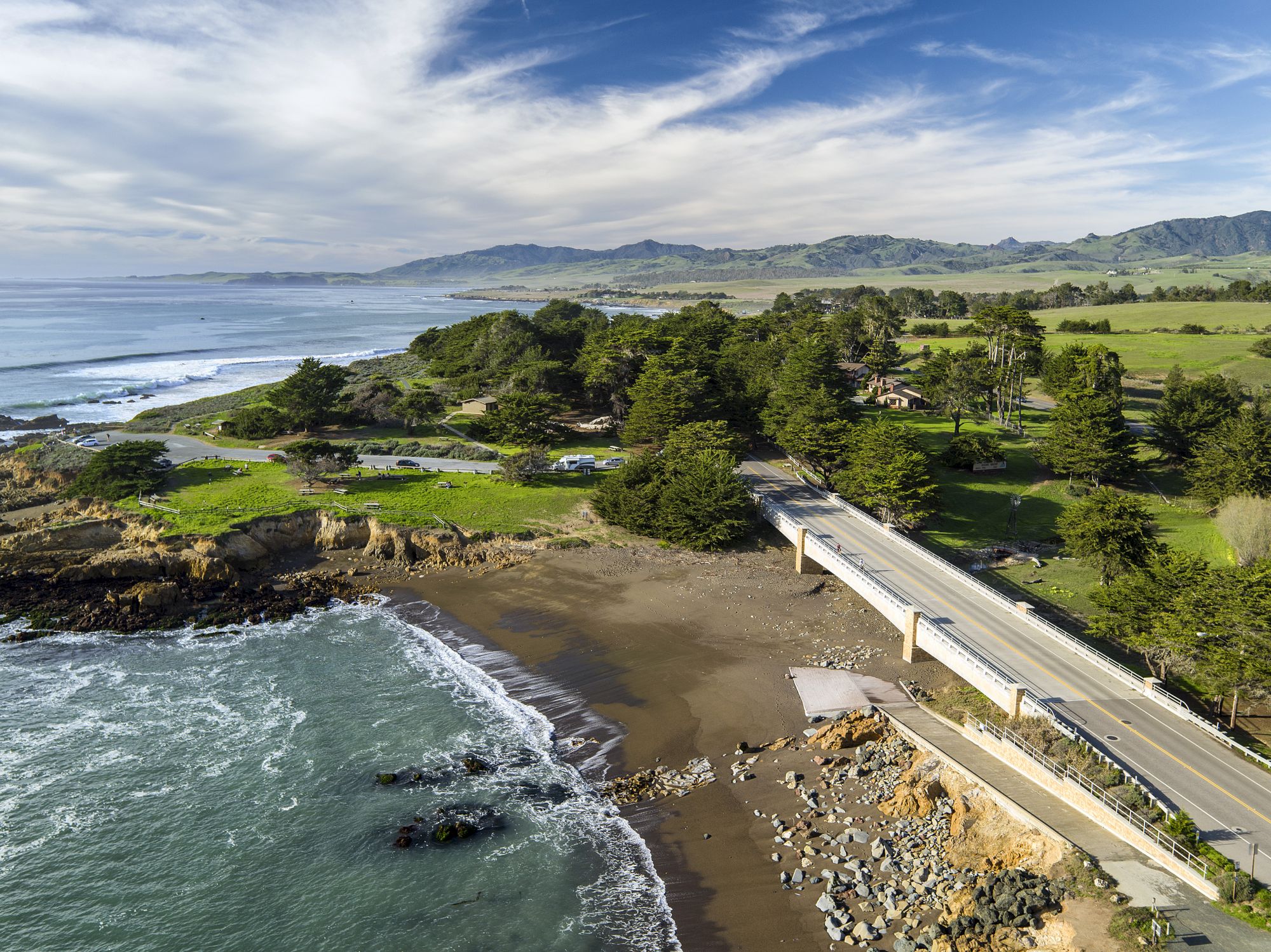 An aerial view shows a coastal beach, rocky shore, and a winding bridge road through lush green hills under a blue sky.
