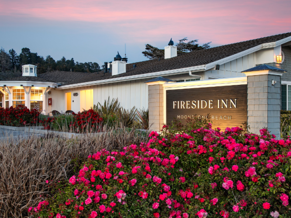 A seaside inn with a welcome sign, pink flowers in bloom, and a pastel sunset sky over the building.