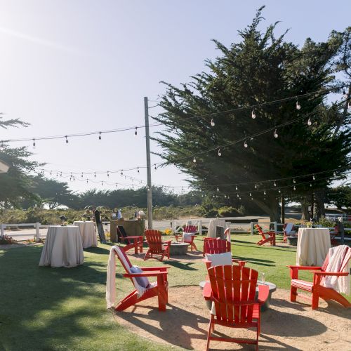 Sunlit outdoor event setup with red benches, white tablecloths, string lights, and large trees in the background, ready for guests.