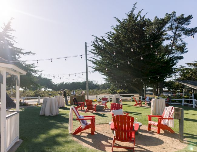 Sunlit outdoor event setup with red benches, white tablecloths, string lights, and large trees in the background, ready for guests.