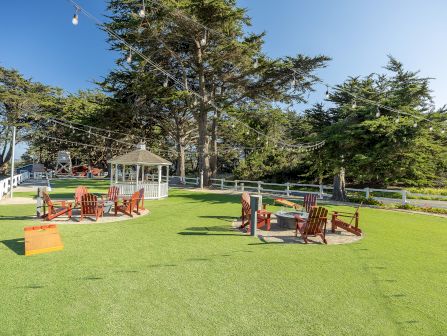 A sunny lawn with orange chairs, a white gazebo, and hanging string lights between tall trees, overlooking a garden.