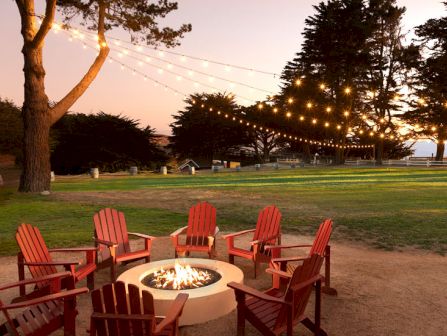 A cozy outdoor fire pit circle with red Adirondack chairs, string lights, trees, and a sunset over a grassy park.
