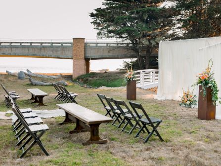An outdoor wedding ceremony setup by a river: rows of chairs, floral bouquet, and a white draped backdrop under a bridge.