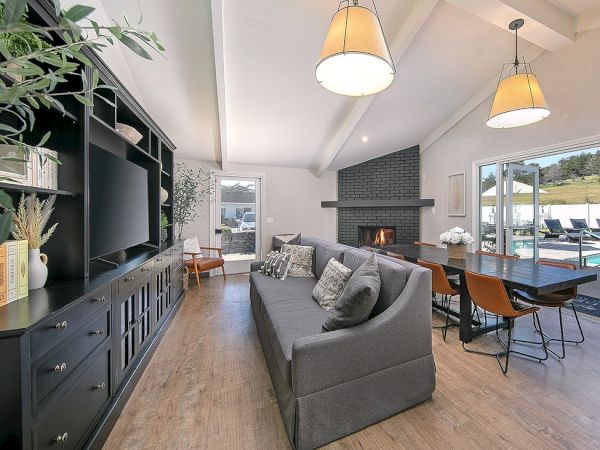 Modern open-concept living room with a gray sofa, built-in media wall, and a cozy dining nook by a brick fireplace, overlooking outdoors.