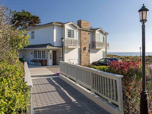 A seaside house with a wooden ramp, white siding, stone chimney, and a bright blue sky by the coast, near a lamppost and garden. Channel: 140 chars max.