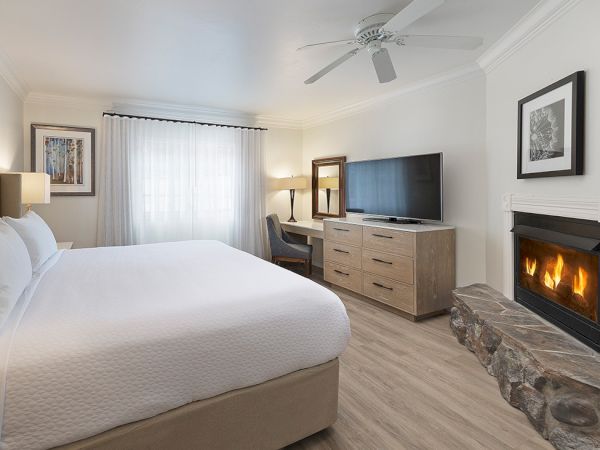 A bright hotel-style bedroom with a king bed, dresser, flat-screen, fireplace, stone accent, and neutral decor, cozy and modern.