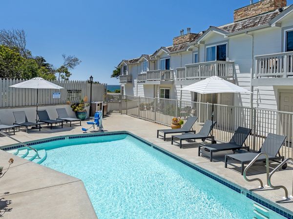 A bright outdoor pool area with clear water, lounge chairs, umbrellas, and a row of light apartment buildings in the background, sunny day.