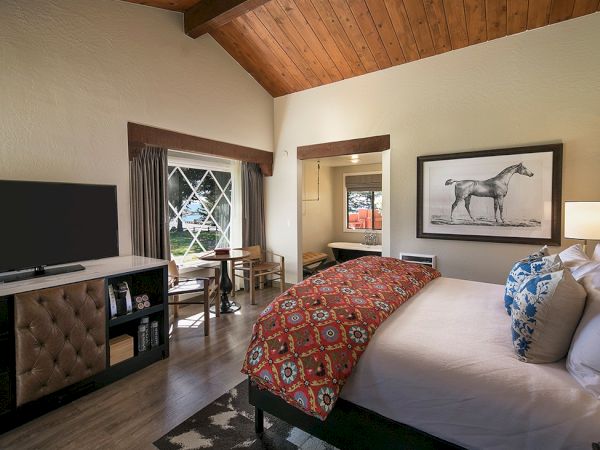A cozy hotel bedroom with a wooden vaulted ceiling, king bed, desk, seating by a window, a dresser with TV, and horse artwork above the bed.