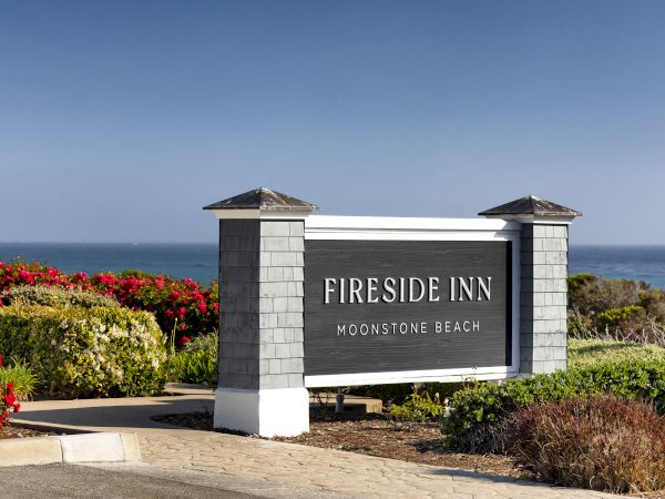 A roadside sign reads &ldquo;Fireside Inn Moonstone Beach&rdquo; with the ocean in the background and nearby red flowers.