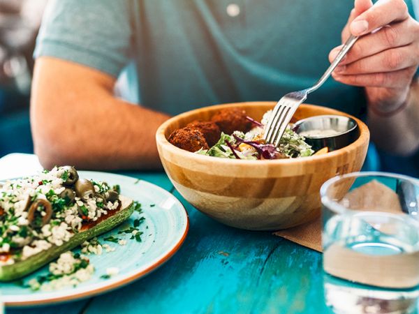 A person eats a wooden bowl meal with a fork while a plate of greens and quinoa sits nearby, and a glass of water rests on a blue table.