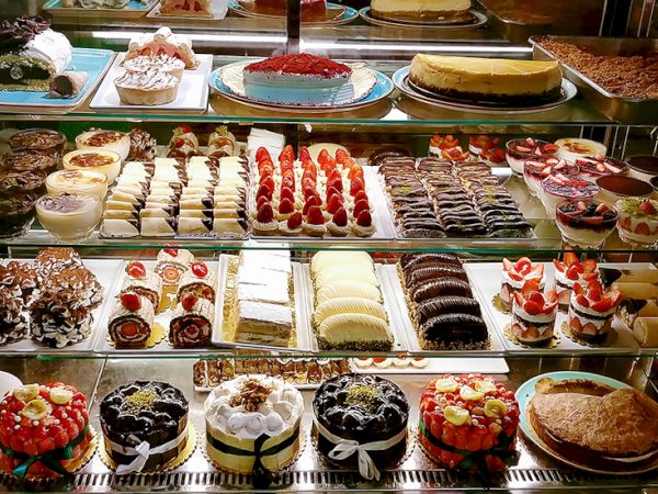Assorted pastries and cakes in a bakery display: mini tarts, cream puffs, eclairs, slice cakes, and chocolate desserts, all arranged neatly.