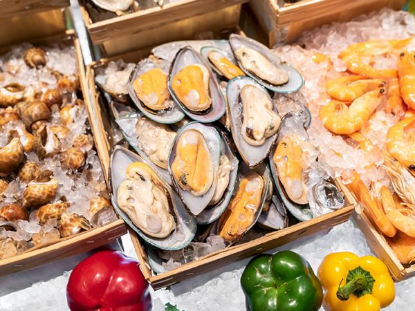 A tray of fresh oysters on ice, with shells open and orange roe visible, surrounded by other shellfish and colorful bell peppers.