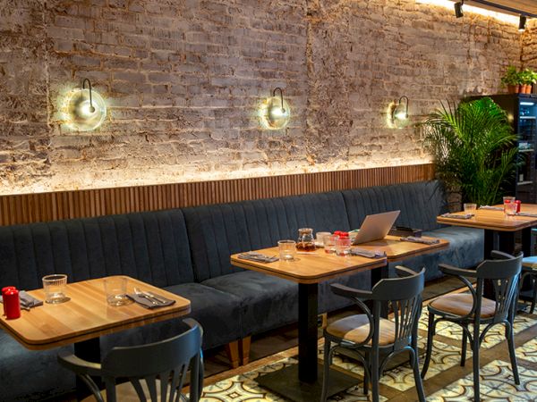 Cozy cafe with brick wall, blue cushioned banquette, wooden tables, metal chairs, wall lights, potted plant, and laptops/menus on tables.