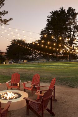 A cozy outdoor fire pit with red Adirondack chairs sits under string lights at sunset, surrounded by trees and a grassy lawn.