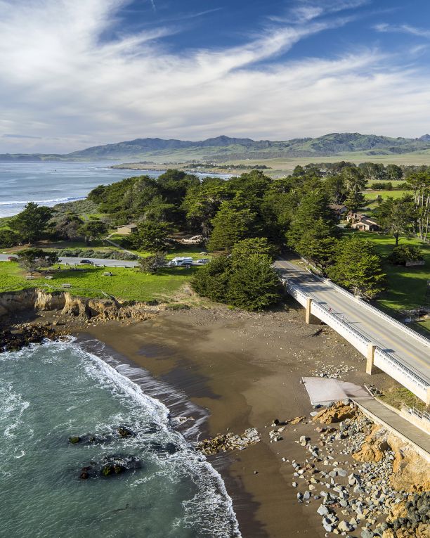 Aerial view of a scenic coastline with a sandy beach, waves, lush green hills, and a road bridge along the shore.