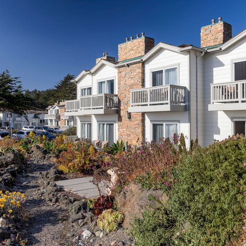 A seaside hotel with white-and-brick buildings, balconies, landscaped gardens, and a &ldquo;Pelican Inn & Suites&rdquo; sign under a blue sky.