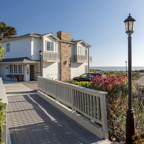 A seaside home with white balcony walkway, garden flowers, a black lantern post, and a car parked near the ocean under blue sky.