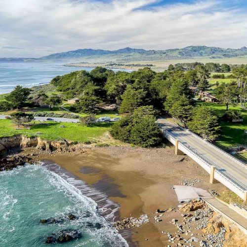 A scenic coastline with a sandy beach, green countryside, trees, a small bridge/road, and distant hills under a blue sky.