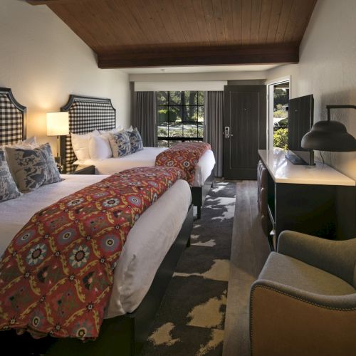A hotel-style bedroom with two beds, patterned blankets, a lamp, curtains, a window view, and a desk with chair.