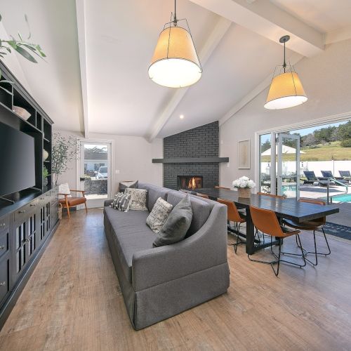 A bright, modern living room with a gray sofa, built-in dark shelving, a fireplace, and a dining area by large windows overlooking a pool and hillside.