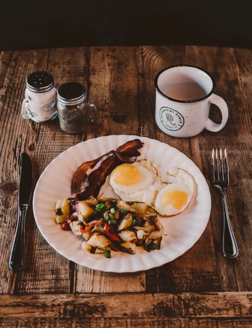 Breakfast on a wooden table: two sunny-side eggs, bacon and potatoes, a mug of coffee, and salt/pepper shakers.