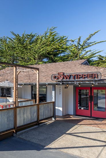 An outdoor patio with tables and umbrellas beside a building entrance labeled &ldquo;TWIN TIPPERS&rdquo; under a clear blue sky.