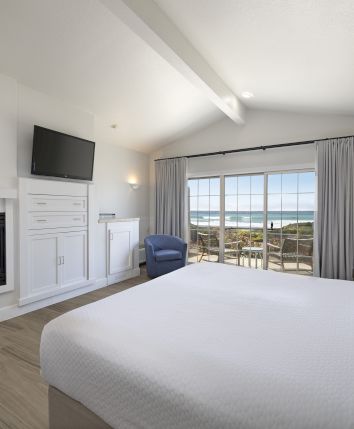 A bright coastal bedroom with a lit fireplace, flat-screen TV, and ocean view through sliding glass doors.