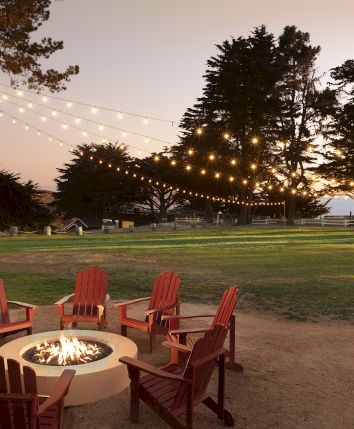 A cozy firepit scene with red Adirondack chairs, warm string lights, and tall trees at sunset in an outdoor campground.