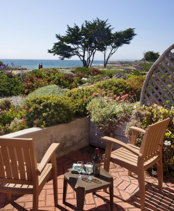 A sunny garden patio with two chairs, wine on a table, colorful flowers, and an ocean view under a clear blue sky.