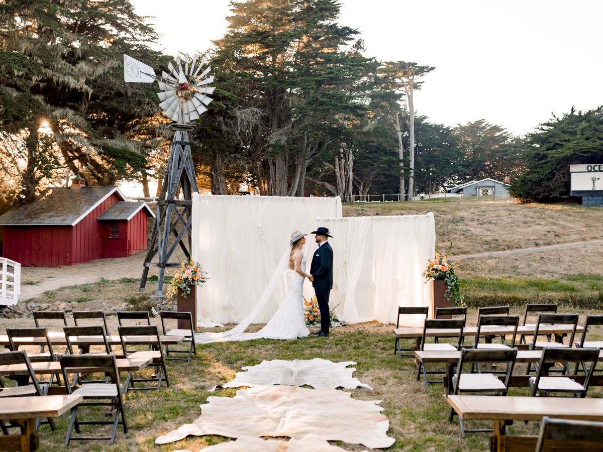 An outdoor wedding ceremony: a bride and groom stand by a white draped arch, with empty chairs, a windmill backdrop, and trees.