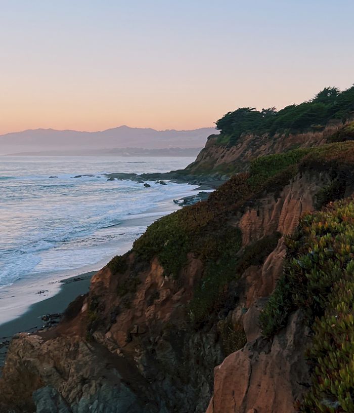 Coastal cliffside at sunset, waves crash on a rocky shore, evergreen brush covers the hillside, warm sky fades over distant hills.