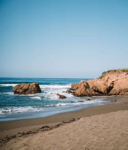 A sunny shoreline with rolling ocean waves, rocky outcrops, and sandy beach along a low coastal cliff under a clear blue sky.
