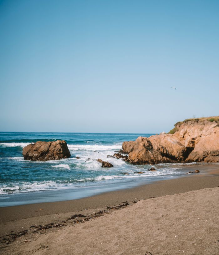 A sunny shoreline with rolling ocean waves, rocky outcrops, and sandy beach along a low coastal cliff under a clear blue sky.