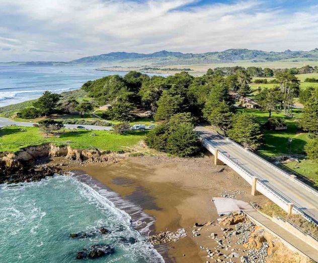 A coastal park with a small sandy beach, rocky shoreline, pine trees, and a bridge crossing to a grassy area by the sea, under blue skies.