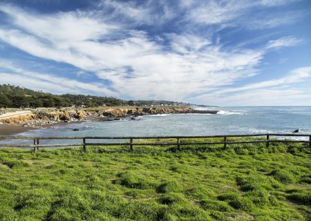 A grassy coastal scene with a fence along the shore, rocky outcrops by the water, and a bright blue sky scattered with white clouds.