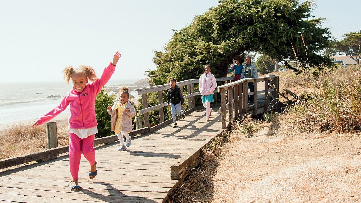 A child in a pink outfit runs happily along a wooden coastal boardwalk with other kids by the ocean.