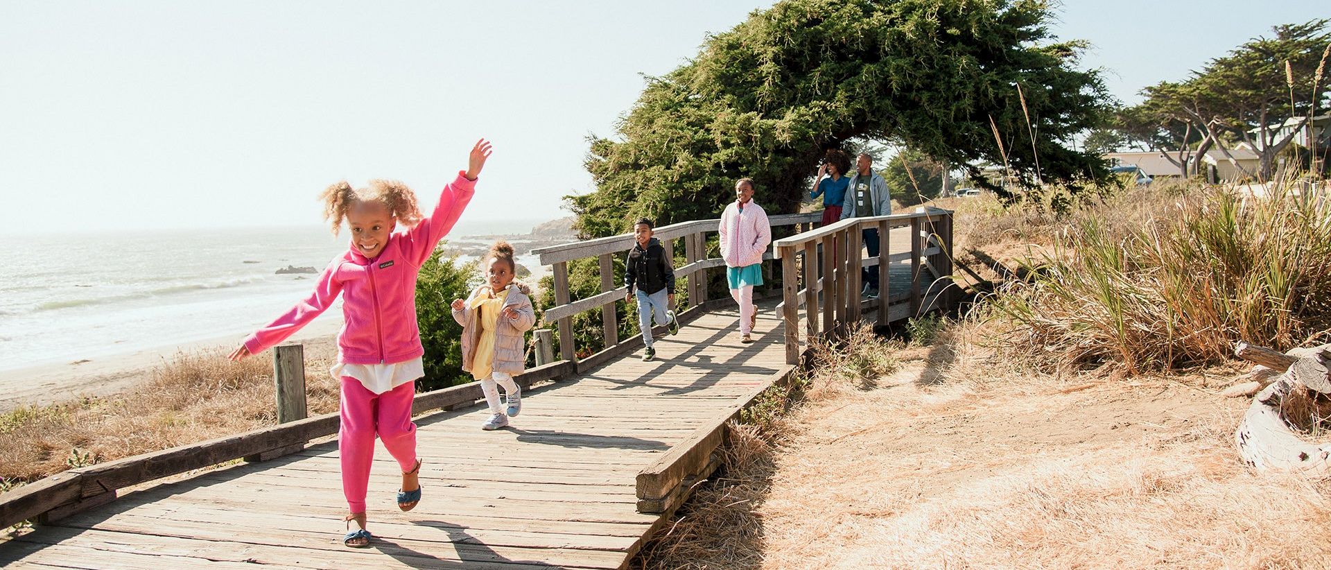 A child in a pink outfit runs happily along a wooden coastal boardwalk with other kids by the ocean.