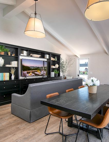 A modern dining and living room featuring a dark table with leather chairs, built-in shelving, plants, and ceiling pendant lights.