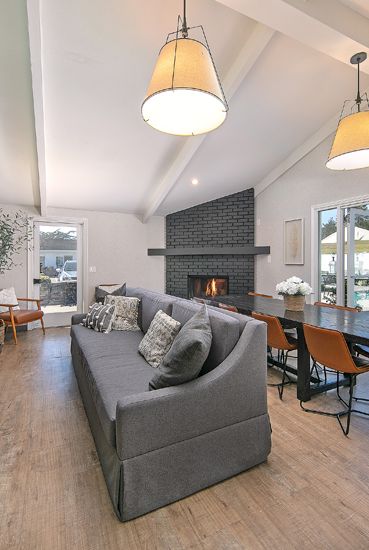 Bright modern living room with a gray sectional, fireplace, dining table, large windows, and an outdoor view.